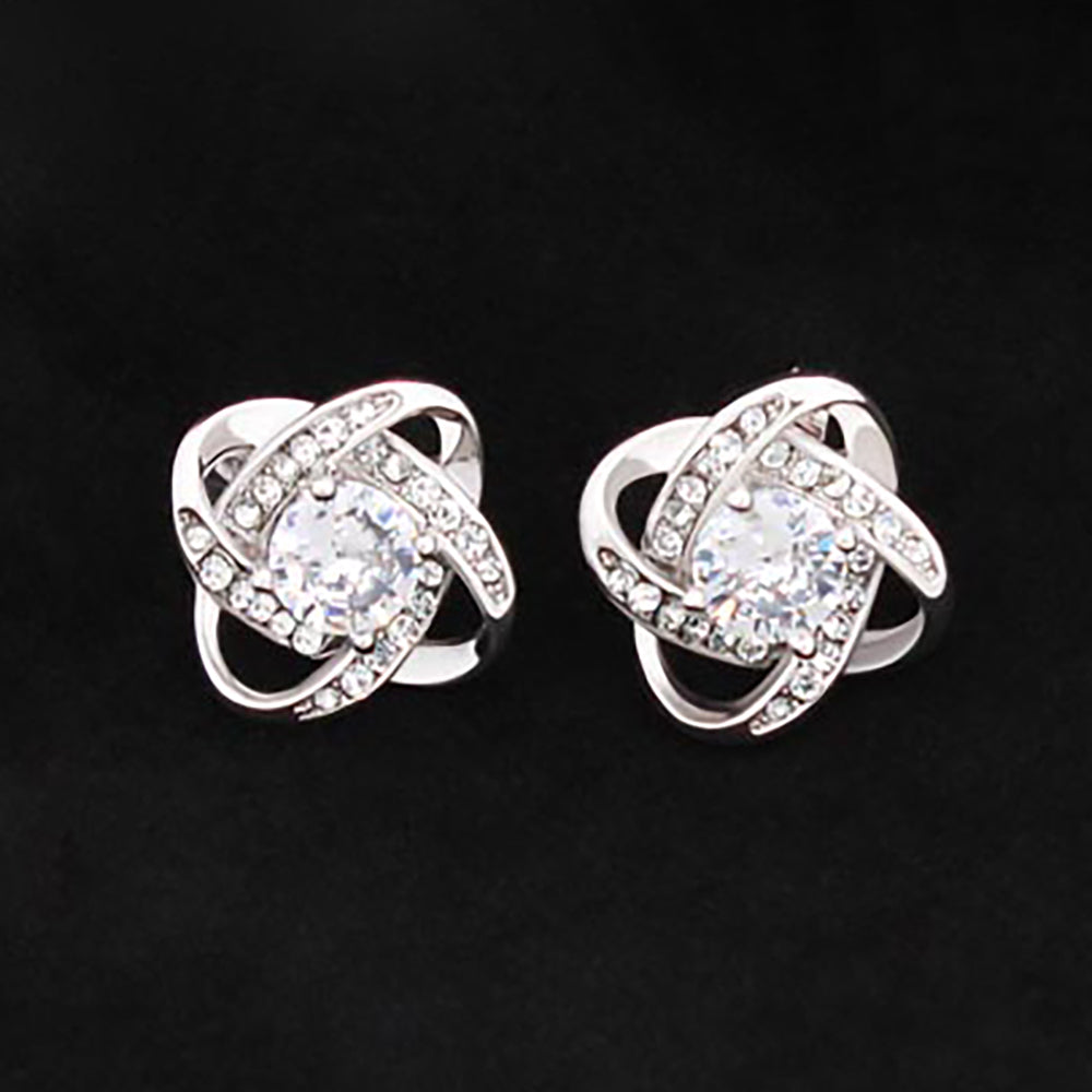 A pair of love knot stud earrings with a 14K white gold over stainless steel, featuring a central cubic zirconia with smaller clear stones around it, displayed against a black background.